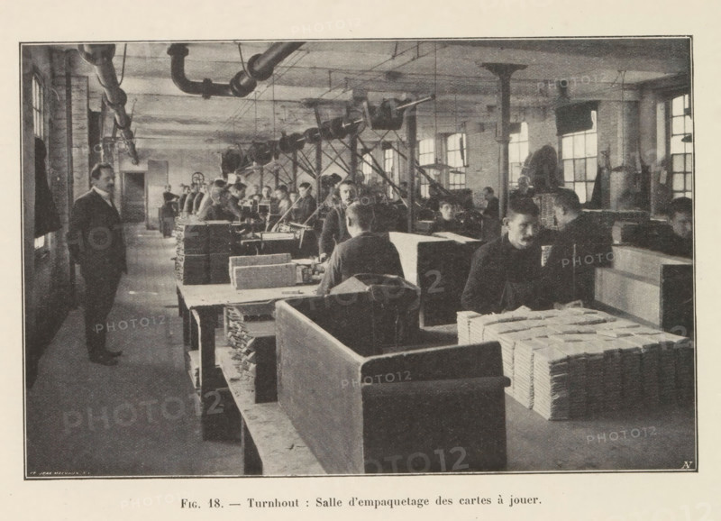 Men pack printed playing cards in a printing house in Turnhout, page, photomechanical print, monogram: 'AN', inscription: 'Fig. 18. - Turnhout: Salle d'empaquetage des cartes à jouer.', photographer: unknown, Turnhout, cliché maker: Jean Malvaux, c. 1901 - in or before 1906, unknown, Jean Malvaux, photographer, cliché maker, paper, autotypie, height 110 mm x width 170 mm, printing, printing office, adult man, playing-cards, workshop-interior, A wide interior scene shows a long room with rows of workbenches and stacks of rectangular bundles on and beside the benches. Men in dark clothing are seated and standing along the benches, some facing the camera and some turned toward the work, while a few figures stand in the aisle. Overhead pipes and a system of pulleys or belts run along the ceiling supported by columns, and light comes through tall multi-pane windows along the far wall. In the foreground there are large wooden partitions and neatly piled bundles on the benches, and the overal...