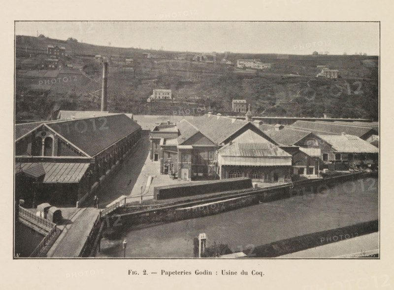 View of a building of the Godin paper factory in Huy, page, photomechanical print, inscription: 'Fig. 2. - Papeteries Godin: Usine du Coq', monogram: 'AN', photographer: unknown, Huy, cliché maker: Jean Malvaux, c. 1901 - in or before 1906, Hoei, paper, autotypie, height 110 mm x width 171 mm, factory-building, A sepia-toned aerial view of an industrial complex with long brick buildings and pitched roofs occupying the foreground, including a large structure with arched windows and a smaller building with a metal roof and decorative cupola, adjacent courtyards and paved walkways, a bridge leading into the site with a small vehicle and a lone pedestrian, a tall smokestack rising near the center, a stone wall separating the complex from an open water channel in front, and rolling hills in the background dotted with scattered houses and small farm buildings under a light sky.