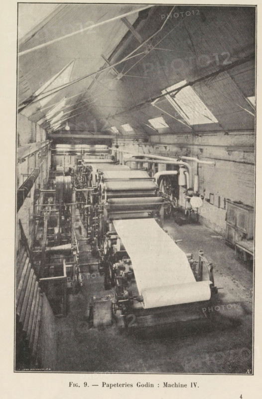 View of a paper production machine at the Godin paper mill in Huy, page, photomechanical print, monogram: 'AN', inscription: 'Fig. 9. - Papeteries Godin: Machine IV.', photographer: unknown, Huy, cliché maker: Jean Malvaux, c. 1901 - in or before 1906, Hoei, paper, autotypie, height 171 mm x width 110 mm, workshop interior, machine driven by electricity, A high-ceilinged industrial room with rows of large cylindrical rollers and machinery aligned along the floor, a wide sheet of material fed across the rollers, overhead skylights set into a sloped roof with visible supports and rigging, exposed pipes and ducts running along the walls, a concrete or worn floor, ladders and platforms adjacent to the machinery, and a few mechanical components and cabinets positioned against the side walls.