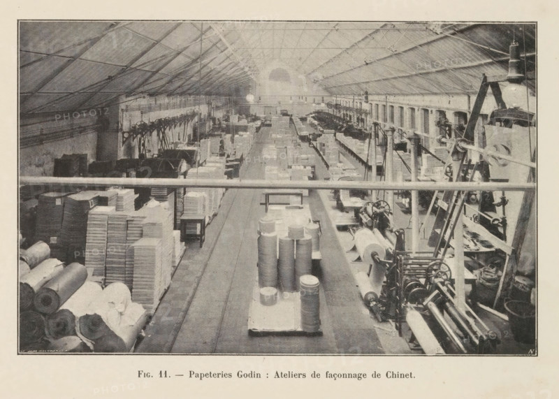 View of a workshop in the Godin paper factory in Huy, page, photomechanical print, monogram: 'AN', inscription: 'Fig. 11. - Papeteries Godin: Ateliers de façonnage de Chinet.', photographer: unknown, Huy, cliché maker: Jean Malvaux, c. 1901 - in or before 1906, Hoei, paper, autotypie, height 110 mm x width 171 mm, workshop interior, A wide factory interior viewed from an elevated perspective shows a long central aisle flanked by worktables, stacks of cylindrical rolls and rectangular bundles, and rows of machinery with visible gears, spools, and rollers; the high pitched roof has a lattice of beams and hanging lamps, and light enters from a large arched window at the far end; on the left side there are piles of rolled material and stacked boxes, while the right side features mechanical equipment, ladders, and more spools and levers aligned along the wall; structural columns and railings run along the foreground and sides, and the overall scene is monochrome with textured shading.