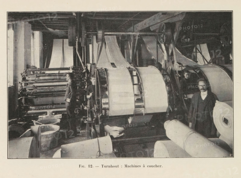View of a workshop in a paper factory in Turnhout, page, photomechanical print, monogram: 'AN', inscription: 'Fig. 12. - Turnhout: Machines à coucher.', photographer: unknown, Turnhout, cliché maker: Jean Malvaux, c. 1901 - in or before 1906, paper, autotypie, height 110 mm x width 169 mm, workshop interior, A monochrome photograph shows an industrial interior with large rolling machines and wide rolls of material. Several vertical and horizontal shafts, gears, and rollers occupy the central area, with sheets of material running over and between them. Men stand among the machinery; one man is positioned near the foreground beside a large roll, another is partially visible behind a machine, and another stands further back by the rollers. Wooden beams and structural supports are visible overhead, and buckets or tubs sit on the floor near the machines. The image is framed by a light border and a lighter margin surrounds the photograph.