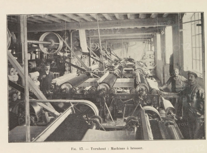 View of a workshop with brushing machines in a paper factory in Turnhout, page, photomechanical print, inscription: 'Fig. 13. - Turnhout: Machines à brosser.', monogram: 'AN', photographer: unknown, Turnhout, cliché maker: Jean Malvaux, c. 1901 - in or before 1906, paper, autotypie, height 110 mm x width 170 mm, workshop interior, machine driven by electricity, A wide interior view of a workshop filled with rows of large mechanical rollers and interconnected belts and pulleys arranged along the length of the room, with stacks of cylindrical rolls of material visible on shelves and along the sides, wooden ceiling beams overhead and vertical support posts, a long window letting light in from one side, and several workers standing near the machinery wearing work clothes and aprons.