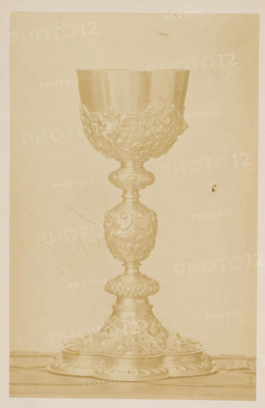 Chalice, photograph, page, c. 1870 - in or before 1875, photographer, paper, albumen print, height 189 mm x width 121 mm, A tall ornate chalice with a wide bowl and a detailed, carved stem stands centered on a flat surface, shown against a plain, light background; the bowl and stem feature floral and scrollwork reliefs and rounded decorative nodes, and the base spreads outward with layered circular platforms and more ornamental relief; the image has a sepia tone and shows faint scratches and small blemishes on the background.