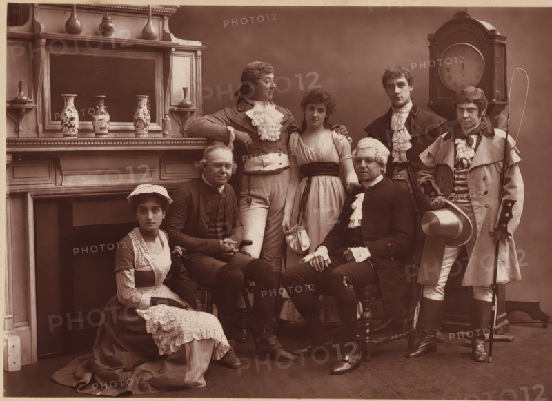 Group portrait of the members of The Vaudeville Company in 'The Road to Ruin', page, photomechanical print, photographer: Herbert Rose Barraud, c. 1881 - in or before 1886, paper, Woodburytype, height 114 mm x width 164 mm, Print two pages before page 178., Person, Kate Rorke, Charles Warner, James Fernandez, Thomas Thorne, Fuller Mellish, Fred Thorne, Louisa Peach, historical persons, adult woman, adult man, A group of seven people posed indoors in period clothing, arranged in two seated and five standing positions; the setting includes a mantelpiece with decorative vases and a large clock behind the group. The seated figures include a woman on the floor wearing a dark dress with a light apron and lace cap, and a man in dark formal attire with a wig and lace cuffs sitting on a carved wooden chair. Standing figures include a man in a light waistcoat and ruffled cravat leaning on the mantel, a young woman in a light gown with a dark sash standing beside him, a man in a dark coat with a ...
