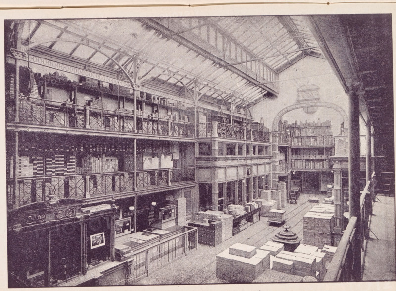 View of a Lutkie & Smit paper warehouse in Amsterdam, Lutkie & Smit Paper Warehouse in Amsterdam, page, photomechanical print, name: 'R...', cliché maker: unknown, c. 1887 - in or before 1892, photographer, paper, autotype, height 129 mm x width 187 mm, Print in a section of advertisements at the back of a book. Print is part of an advertising message by Lutkie & Smit., large-scale structure for storage, eg: warehouse, depot, silo, Amsterdam, A large interior space with a high arched roof supported by exposed beams and metal trusses, multiple tiers of open balconies with decorative railings lining the sides, tall columns rising between levels, stacked crates and boxes arranged on the floor, counters and shelving along the lower level, staircases and walkways connecting the balconies, hanging lamps and circular light fixtures suspended from the ceiling, architectural details such as ornate cornices and carved wood panels, and a curved archway visible at the far end.