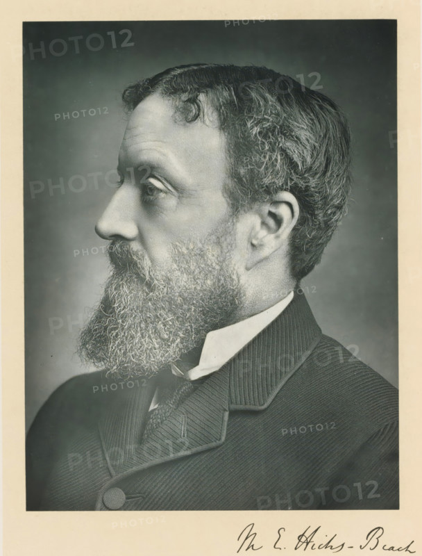 The right honorable sir Michael Edward Hicks-Beach, photograph, page, inscription: '17, Baker St, London, W.', annotation, photographer: Russell & Sons, c. 1893 - c. 1903, paper, carbon print, height 227 mm x width 171 mm, Person, Michael Edward Hicks Beach, historical persons (portraits and scenes from the life) ((full) bust portrait), A black and white portrait showing a close-up profile of a man facing left with a full, curly beard and mustache, short wavy hair with visible texture, and a prominent ear; he is wearing a high-collared shirt, a patterned tie, and a dark, textured jacket with a rounded button and a notched lapel, all set against a plain, softly shaded background with a light border around the image.