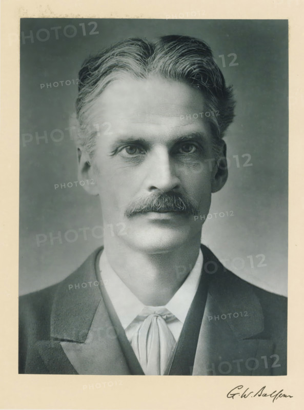 The right honorable Gerald William Balfour, photograph, page, inscription: 'Leeds', annotation, photographer: AC Hoskins, London, c. 1893 - c. 1903, paper, carbon print, height 228 mm x width 170 mm, Person, Gerald Balfour, historical persons (portraits and scenes from the life) ((full) bust portrait), A black and white portrait of a man from the shoulders up centered on a neutral gray background, showing a face with a neatly groomed mustache, high cheekbones, and deep-set eyes looking forward, with wavy hair swept back from a defined part at the top, visible ear on one side, and faint wrinkles on the forehead and around the eyes; he is wearing a dark jacket over a vest and a light-colored shirt with a tied neckcloth, and a light border surrounds the photograph.
