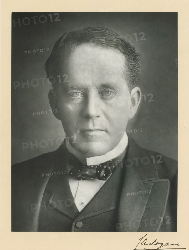 The right honorable earl Cadogan KG, photograph, page, annotation, inscription: 'Dublin', photographer: Chancellor, Ireland, c. 1893 - c. 1903, paper, carbon print, height 228 mm x width 170 mm, Person, George Cadogan, historical persons (portraits and scenes from the life) ((full) bust portrait), A black and white portrait of an older man from the chest up centered in the frame, showing a close-up of his face with neatly combed hair, pronounced forehead, and neutral expression, his eyes looking forward and visible under subtle shadows, ears partially visible, and a smooth complexion with fine facial lines; he is wearing a dark suit jacket over a waistcoat and a white high collar, with a patterned bow tie at the throat; the photograph has a narrow light border and a darker background behind the man.