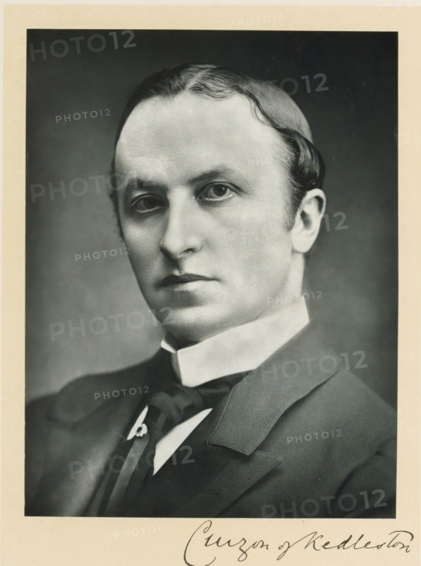 The right honorable lord Curzon of Kedleston, photograph, page, inscription: '55, Baker St, London, W.', annotation, photographer: Elliott & Fry, c. 1893 - c. 1903, paper, carbon print, height 227 mm x width 171 mm, Person, George Curzon, historical persons (portraits and scenes from the life) ((full) bust portrait), A black and white portrait of a man shown from the shoulders up wearing a dark suit jacket, a high white collar, and a dark tie with a decorative pin. He has a smooth, pale complexion, neatly combed hair with a waved part, and an expressionless face with slightly downturned lips. The background is plain and darker than his face, and a narrow light-colored border surrounds the photograph.