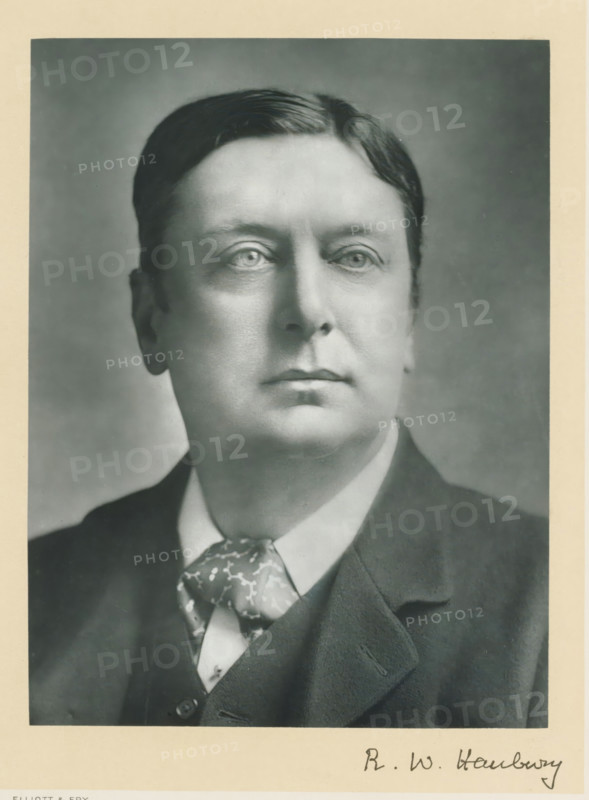 The right honorable Robert William Hanbury, photograph, page, annotation, inscription: '55, Baker St, London, W.', photographer: Elliott & Fry, c. 1893 - c. 1903, paper, carbon print, height 227 mm x width 171 mm, Person, Robert William Hanbury, historical persons (portraits and scenes from the life) ((full) bust portrait), A black and white portrait photograph of a man from the shoulders up, showing a close-up of his face with short, neatly combed hair, light-toned skin, and a calm expression. He is wearing a dark suit jacket over a vest and a white shirt, with a patterned necktie visible at the collar. The background is a plain, softly shaded backdrop, and the image has a narrow light-colored border around it.