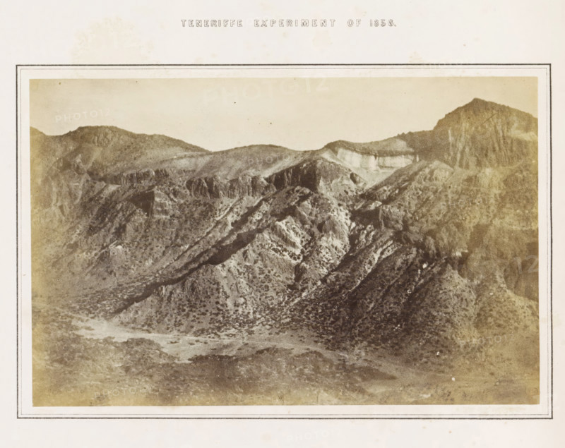 Tenerife Experiment of 1856, View of the crater of El Teide on Tenerife, photograph, page, number: 'Plate 3.', inscription: 'Portion of a Photograph (magnified 9.1 diameters), representing a mountain side (the interior S. Eastern wall of the Great Crater), four miles distant from the Camera, stationed at a height of 10, 700 feet above the level of the Sea. See pp 513, 514.', photographer: Charles Piazzi Smyth, Tenerife, printer (person): JA Spencer (possibly), 1856, printer (person), paper, albumen print, height 128 mm x width 197 mm, Photo in the section at the back., experiment, test, science and technology, volcano, A sepia-toned photograph of a rugged mountain landscape framed by a pale border, showing layered ridges and steep rocky slopes with scattered low vegetation, terraces and cliffs visible along the higher slopes, a broad valley floor with uneven ground and textured scrub, and distant rounded peaks beneath a pale sky.