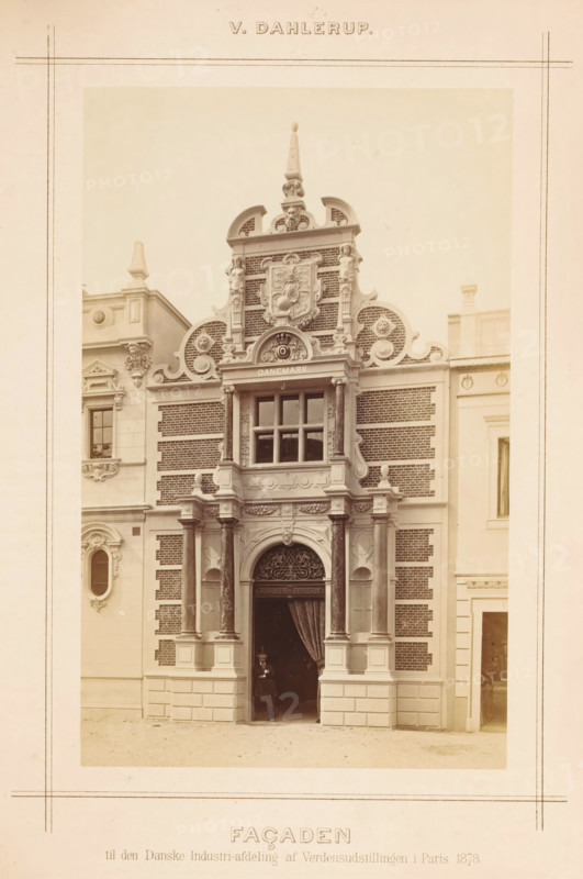 View of a Danish-style façade at the Paris World Exhibition in 1878, designed by Vilhelm Dahlerup, Façaden, photograph, page, inscription: 'til den Danske Industri-afdeling af Verdensudstillingen i Paris 1878.', photographer: Johannes Jaeger, 1878-05-01 - 1878-11-10, paper, albumen print, albumendruk, height 201 mm x width 126 mm, front façade, Wereldtentoonstelling 1878 (Paris), Paris, Period, A sepia-toned photograph of an ornate building facade framed by a pale border, showing a central arched entrance flanked by paired columns with decorative capitals and bases, above which is an elaborate sculpted pediment with scrollwork, circular motifs, and a carved coat of arms set into a patterned brick background; a rectangular window with muntins sits above the entrance within a projecting bay supported by pilasters and intricate molding, while balustrade-like details and small finials decorate the upper sections; the facade combines smooth stone blocks at the lower level with darker textur...