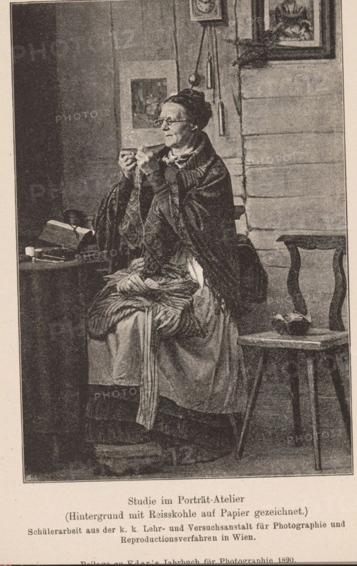 Studio portrait of an unknown seamstress, page, photomechanical print, inscription: 'Studio im Portät-Atelier, (Hintergrund mit Reisskohle auf Papier gezeichnet.)', maker: KK Lehr- und Versuchsanstalt für Photographie, c. 1885 - in or before 1890, paper, autotypie, height 138 mm x width 95 mm, Print on the twenty-first unnumbered page after page 400., historical persons - BB - woman, sewing, An older woman sits on a bench wearing layered garments and a shawl, holding a small object close to her face with both hands and looking slightly to the side, her hair pulled back and she wears glasses; beside her a small table holds a book and other objects, and a simple wooden chair stands nearby with a bowl-like item on its seat; the background shows a wall with framed pictures and hanging objects, and the scene is rendered in a monochrome, stippled or engraved style.