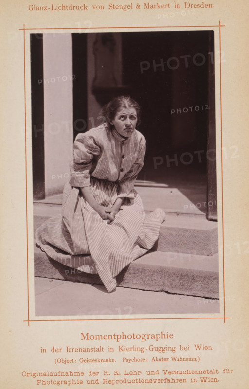 Portrait of an unknown woman in a psychiatric institution, page, photomechanical print, inscription: 'Momentphotographie, in der Irrenanstalt in Kierling-Gugging bei Wien., (Object: Geisteskranke. Psychose: Akuter Wahnsinn.)', photographer: KK Lehr- und Versuchsanstalt für Photographie, Klosterneuburg, cliché maker: Stengel & Markert, Dresden, c. 1885 - in or before 1890, Lower Austria, Saxony, paper, collotype, photoprint, height 131 mm x width 85 mm, Print on fifth unnumbered page after page 400., historical persons - BB - woman, diseases, A monochrome photograph shows a woman seated on stone steps in front of a dark doorway. She wears a light-colored, striped dress with long sleeves and buttons down the front, and the skirt is gathered around her on the step. Her hands are clasped together in her lap and her body leans slightly forward. Her hair is pulled back and appears slightly disheveled. The doorway behind her is shadowed, with a lighter vertical wall panel visible to one side....