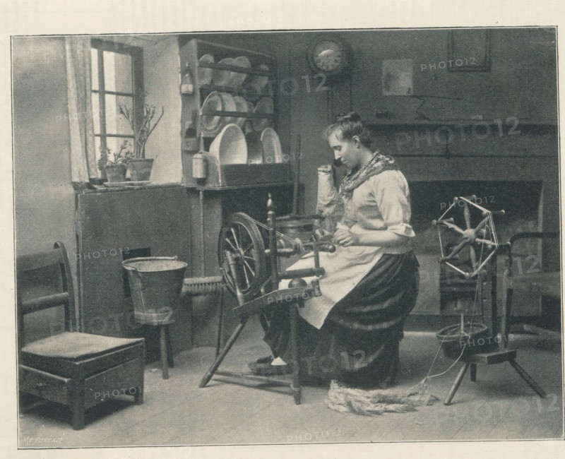 Woman at spinning wheel, Spinning, page, photomechanical print, photographer: John William Anckorn, cliché maker: Meisenbach's Autotype Company, c. 1889 - in or before 1894, John William Anckorn, Meisenbach's Autotype Company, paper, autotypie, height 114 mm x width 151 mm, spinning-wheel, A monochrome scene showing a woman seated at a spinning wheel, wearing a light blouse with rolled sleeves, a patterned scarf around her shoulders, and a dark skirt with an apron; she is focused on her hands near the wheel mechanism and a bundle of fiber lies on the floor beside her. The room contains wooden furniture including a low box-like stool, chairs, and a shelf unit holding plates and bowls; a potted plant sits on the windowsill beside a curtained window. In the background there is a wall clock and a mantel with framed items above it, and a metal bucket rests on a small stand near the furniture. The image has a textured, halftone appearance and is bordered by a narrow frame.