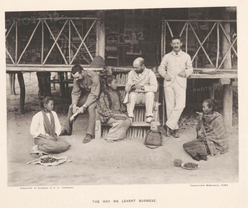 The way we learned Burmese, Three men with books and three women from Burma in front of a house, page, photomechanical print, photographer: AGE Newland, Burma, maker: Michel Berthaud, Paris, c. 1892, paper, collotype, photoprint, height 154 mm x width 202 mm, Prints opposite page 20., book, hut, cabin, lodge, A group of people sits and kneels in front of a raised wooden structure with a woven bamboo railing and vertical posts; two men in light-colored suits sit or stand near the railing, one seated reading from a small book and the other standing with a hand resting on the railing next to a hat, while another man in a darker uniform leans forward showing a piece of paper to a seated woman in patterned clothing who holds a small bundle in her lap; two other women kneel on the ground at either side, one with a scarf around her neck and a cloth of small round objects spread before her and the other wrapped in a patterned shawl holding a small item near her mouth with plates of small round...
