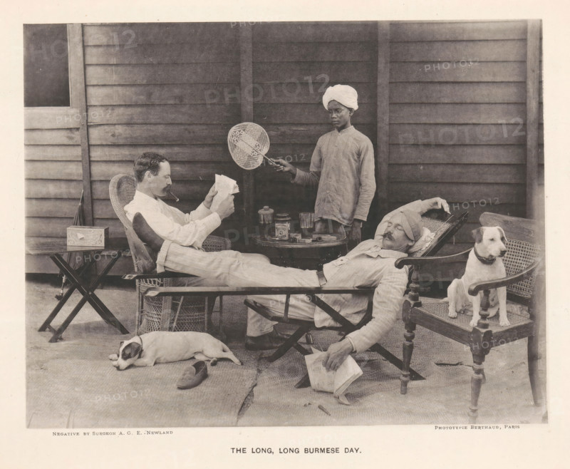 Two seated men and a boy with a fan behind them, The long, long Burmese day, page, photomechanical print, photographer: AGE Newland, Burma, maker: Michel Berthaud, Paris, c. 1892, paper, collotype, photoprint, height 157 mm x width 202 mm, dog, reading, fan, A monochrome scene shows a wooden paneled wall as a backdrop with three men and two dogs arranged on a porch. One man reclines in a wicker chair wearing light clothing and a turban, his legs stretched out and a pipe in his mouth while he reads a folded paper; another man reclines on a lounge with his head wrapped and eyes closed, one arm resting behind his head and the other hand holding a book; a standing man in a buttoned shirt and a turban holds a round woven fan on a handle over the seated men. A small round table between the chairs holds glassware and a covered jar. Two dogs are present, one lying on the floor near a shoe and the other sitting alert on a wooden chair beside the reclining man. The floor is covered with mats and...