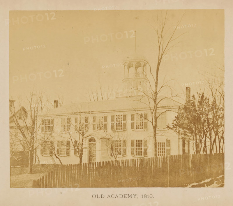 View of the first Friends Academy location in New Bedford, Massachusetts, Old Academy. 1810, photograph, page, c. 1871 - in or before 1876, photographer, paper, albumen print, height 109 mm x width 132 mm, scholastic education, tuition, civic architecture, edifices, dwellings, North America, New Bedford, A faded sepia photograph of a large rectangular building with multiple shuttered windows across two stories, a pitched roof, and a small central doorway, partially obscured by leafless trees in front and a wooden picket fence along the foreground; a cupola with arched openings rises above the roofline and a tall thin pole extends upward nearby, while more trees and a chimney appear on either side, all shown against a pale sky.
