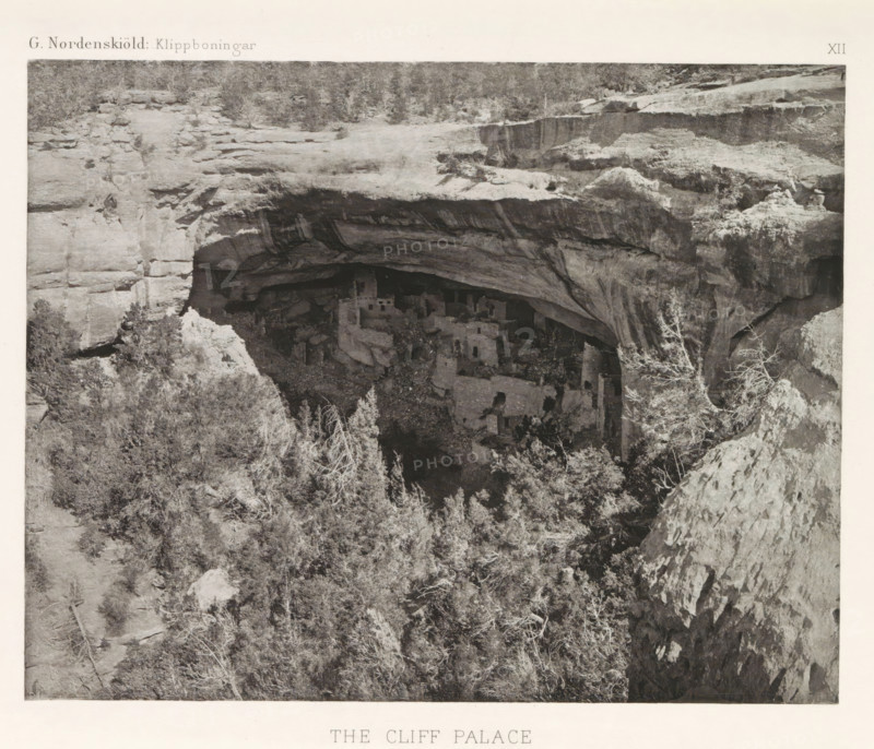 The Cliff Palace, View of the Cliff Palace in Mesa Verde National Park, page, photomechanical print, number: 'XII', inscription: 'G. Nordenskiöld: Klippboningar.', photographer: Gustaf Nordenskiöld, Mesa Verde National Park, cliché maker: Librairies-Imprimeries Réunies (Ancienne Maison Quantin), Paris, c. 1891 - in or before 1893, North America, Colorado, paper, photoaquatint, height 190 mm x width 242 mm, ruin of a dwelling, house, castle, cave-dwelling, Cliff Palace, A wide rock overhang forms a recessed alcove in a cliff face, with layered rock strata and textured surfaces visible on the ceiling and walls of the overhang; beneath the shelter, a cluster of multi-level stone structures with rectangular openings and angular walls sits among scattered boulders and rubble; the foreground shows a slope of rocky terrain and dense vegetation with leafy and twiggy shrubs and small trees, some with pale, bare branches; the upper area of the scene displays a rough plateau of rock with sparse l...