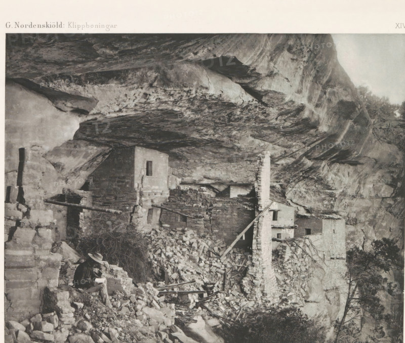 Befästad klippremna ledane till Balcony House, View of an entrance to the Balcony House in Mesa Verde National Park, page, photomechanical print, name, number: 'Fig. 42.', photographer: Gustaf Nordenskiöld (possibly), Mesa Verde National Park, cliché maker: Angerer & Göschl, C., Vienna, c. 1891 - in or before 1893, clichémaker, North America, Lower Austria, Colorado, paper, autotypie, height 201 mm x width 106 mm, ruin of a dwelling, house, castle, cave-dwelling, A grayscale photograph shows a rocky cliff overhang with irregular textured rock surfaces and ledges, beneath which a cluster of stone and adobe structures sits in various states of ruin with exposed walls, a tall narrow chimney, and piles of rubble and scattered masonry; a person wearing a hat and dark clothing is seated on a low stone wall near the rubble holding an open book or object, and wooden beams and planks lean against and span parts of the ruins, with sparse vegetation and small trees visible at the cliff edge.