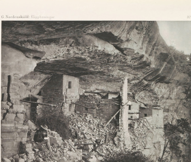 The Balcony House, Balcony House in Mesa Verde National Park, page, photomechanical print, number: 'XIV', inscription: 'G. Nordenskiöld: Klippboningar.', photographer: Gustaf Nordenskiöld, Mesa Verde National Park, cliché maker: Librairies-Imprimeries Réunies (Ancienne Maison Quantin), Paris, c. 1891 - in or before 1893, North America, Colorado, paper, photoaquatint, height 189 mm x width 240 mm, ruin of a dwelling, house, castle, cave-dwelling, A black and white photograph showing a rocky cliff overhang with rough textured rock surfaces and striations, beneath which are multiple stone structures in varying states of ruin with partial walls, narrow rectangular openings, and a freestanding chimney built of stacked stone, piles of loose stones and rubble scattered around, wooden beams and poles leaning against walls and among the debris, sparse vegetation and small trees visible near the cliff edge, and a seated person wearing a hat and darker clothing holding an object while sitting on ...