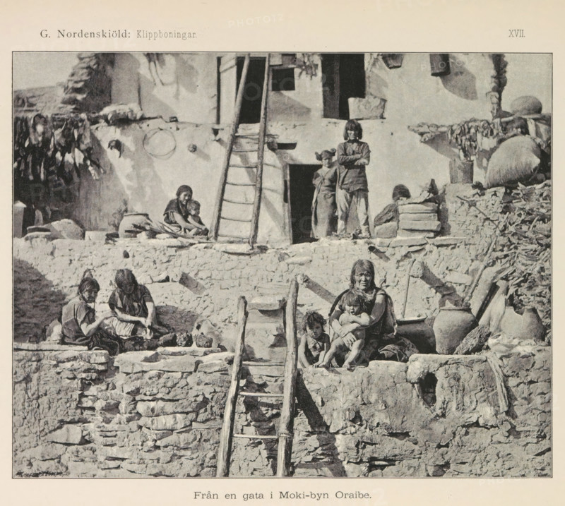 View of a village of the Hopi people, Moki-byn Wol'-pi, page, photomechanical print, name, number: 'Fig. 153.', photographer: Gustaf Nordenskiöld (possibly), North and Central America, cliché maker: Angerer & Göschl, C., Vienna, c. 1891 - in or before 1893, Gustaf Nordenskiöld, Angerer & Göschl, photographer, cliché maker, Lower Austria, paper, autotypie, height 90 mm x width 148 mm, prospect of village, silhouette of village, A grayscale photograph of a stone and plaster building with rough stacked stone walls and a wooden ladder leading up to a doorway, several people sitting and standing on the terraces and steps, a woman holding a child on a ledge with another child nearby, two women seated and working with items in a basin, two figures standing in a doorway on the upper level, pottery jars and bundles placed on the ledges, hanging items draped over the walls, and irregular stone steps and ledges forming the exterior façade.