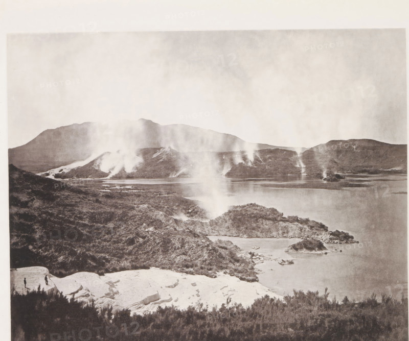 View of Lake Rotomahana and the Pink Terraces, page, photomechanical print, photographer: Daniel Louis Mundy, New Zealand, cliché maker: anonymous, United Kingdom, c. 1865 - c. 1875, paper, collotype, autotype, height 197 mm x width 246 mm, lake, hot spring, geyser, Lake Rotomahana, A wide landscape photograph showing a lake bordered by rocky shorelines and low hills, with steam or smoke rising in several narrow plumes from the ground along the distant shoreline and hills; the foreground includes light-colored rocky outcrops and darker vegetation, and two small human figures standing on a pale rock ledge near the lower left, with distant mountains forming a hazy horizon under a pale sky.