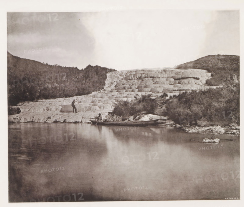 View of the Pink Terraces along Lake Rotomahana, page, photomechanical print, photographer: Daniel Louis Mundy, New Zealand, cliché maker: anonymous, United Kingdom, c. 1865 - c. 1875, paper, collotype, autotype, height 202 mm x width 249 mm, lake, hot spring, geyser, steam, Lake Rotomahana, Pink Terraces, A wide monochrome landscape shows a calm reflective body of water in the foreground with a stepped white terraced formation rising behind it and continuing across the middle of the image, textured with horizontal ledges and small ridges; a wooded hillside frames the terraces on the left and right with darker trees and scrub, and a person stands on one of the lower terraces near the water while another person sits in a small boat at the water’s edge; the sky above is pale with soft cloud patterns and the overall scene has a vintage photographic tone.