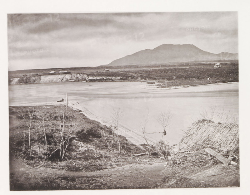 View of Lake Taupo, page, photomechanical print, photographer: Daniel Louis Mundy, New Zealand, cliché maker: anonymous, United Kingdom, c. 1865 - c. 1875, paper, collotype, autotype, height 192 mm x width 250 mm, lake, volcano, hot spring, geyser, Lake Taupo, A wide grayscale landscape showing a calm body of water curving along a sandy shore with a small boat near the waterline, low rocky cliffs and flat terrain behind the shore, scattered leafless trees and rough vegetation in the foreground, a few simple buildings and a solitary small house on the distant plain, telephone or utility poles dotting the scene, and a large rounded mountain range under a cloudy sky.