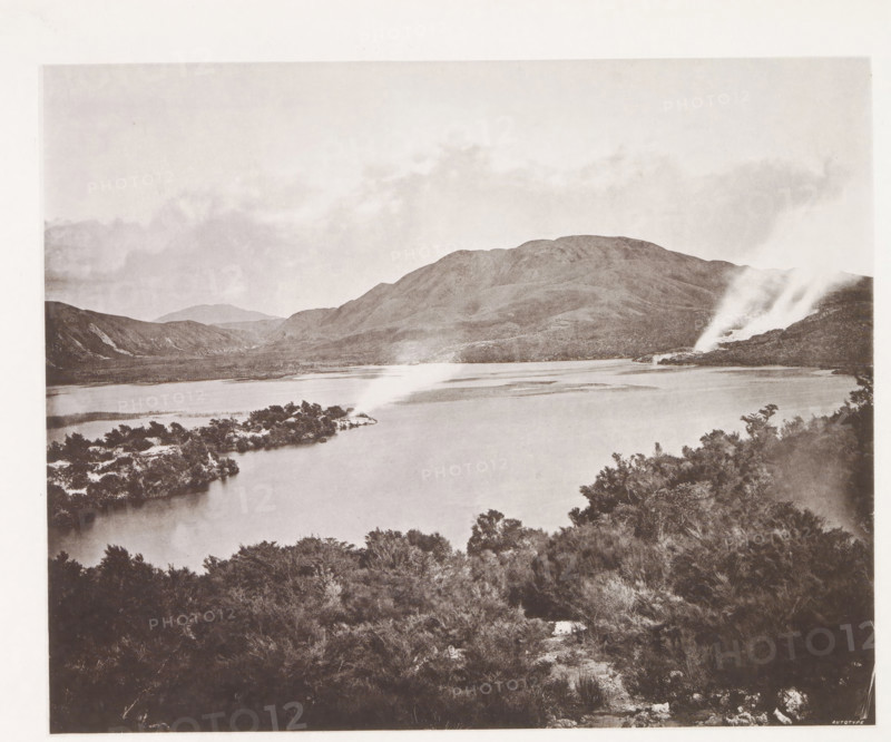 View of Lake Rotomahana, page, photomechanical print, photographer: Daniel Louis Mundy, New Zealand, cliché maker: anonymous, United Kingdom, c. 1865 - c. 1875, paper, collotype, autotype, height 201 mm x width 248 mm, lake, hot spring, geyser, Lake Rotomahana, A wide view of a lake bordered by shoreline vegetation in the foreground and an island with low trees extending into the water, with a range of rounded hills rising behind the lake and a plume of steam or smoke emerging from a rocky area on the right side, under a sky with light cloud coverage.