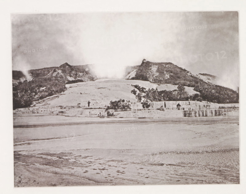 View of the White Terraces at Lake Rotomahana, page, photomechanical print, photographer: Daniel Louis Mundy, New Zealand, cliché maker: anonymous, United Kingdom, c. 1865 - c. 1875, paper, collotype, autotype, height 198 mm x width 245 mm, lake, hot spring, geyser, Lake Rotomahana, White Terraces, A wide sepia-toned landscape showing a layered mineral terrace in the foreground with smooth, rippled surfaces and shallow pools, rising to a stepped shelf where shrubs and small bushes grow, beyond which a rounded slope leads up to a ridge of rocky hills covered with sparse vegetation and patches of lighter exposed rock, a plume of steam or mist rising from the center of the ridge and drifting into a cloudy sky above, and two small human figures standing on the middle terrace near the vegetation.