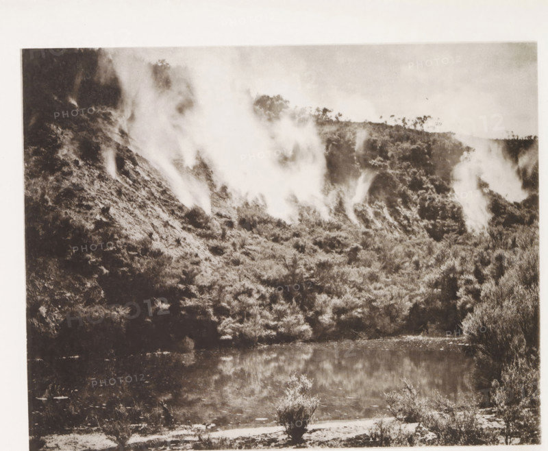 View of steam clouds along Lake Rotomahana, page, photomechanical print, photographer: Daniel Louis Mundy, New Zealand, cliché maker: anonymous, United Kingdom, c. 1865 - c. 1875, paper, collotype, photoprint, height 196 mm x width 249 mm, lake, hot spring, geyser, steam, Lake Rotomahana, A sepia-toned landscape showing a hillside covered in dense vegetation with smoke or mist rising in several plumes along the slope, a band of trees or shrubs along the ridge, a reflective body of water at the base of the hill mirroring the vegetation, and low plants and grassy ground in the foreground.