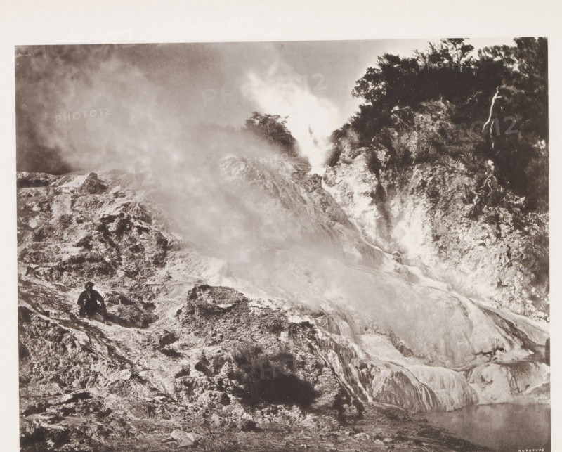 Geyser near Lake Rotomahana, page, photomechanical print, photographer: Daniel Louis Mundy, New Zealand, cliché maker: anonymous, United Kingdom, c. 1865 - c. 1875, paper, collotype, autotype, height 198 mm x width 253 mm, lake, hot spring, geyser, steam, Lake Rotomahana, A black-and-white photograph showing a rocky landscape with steam or mist rising from the ground and flowing across the scene, rough textured terraces and mineral deposits forming layered slopes and ledges, a darker forested area with dense trees and vegetation on the upper right, a solitary figure wearing a hat and dark clothing seated on a rocky outcrop near the lower left, and a pool of water partially visible at the lower right edge.