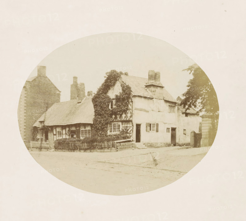 View of a house in Tarvin, Cottage in Tarvin, photograph, page, photographer: William Bryans, Tarvin, c. 1850 - in or before 1858, Cheshire (county), paper, albumen print, height 141 mm x width 175 mm, cottage, A circular sepia photograph shows an old cottage with a thatched roof and timber-framed walls, part of the facade covered in climbing ivy, multiple windows with small panes and shutters, an open doorway, a low wooden fence in front, a stone wall and steps leading up to the entrance, a tall brick chimney and additional brick buildings adjacent, a tree partially visible on one side, and an unpaved road or courtyard in the foreground.