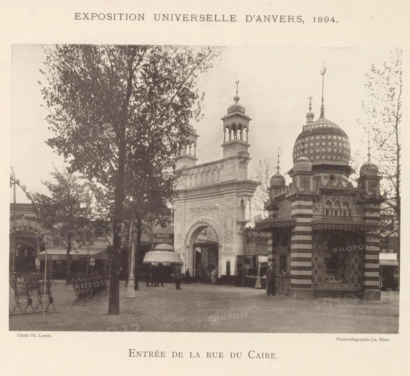 Entrée de la Rue du Caire, Exterieur van het Egypte paviljoen op de wereldtentoonstelling te Antwerpen, page, photomechanical print, photographer: Th. Lantin, Antwerp, printer (person): Joseph Maes, 1894, printer (person), paper, collotype, photogravure, height 160 mm x width 220 mm, façade (of house or building), world fair, world exhibition, Wereldtentoonstelling 1894 (Antwerp), A sepia-toned street scene shows a large ornate archway flanked by decorative towers and a domed pavilion with a patterned surface, finials, and striped lower walls, with trees partially obscuring portions of the architecture and benches arranged in rows in the foreground, several people standing near the arch and pavilion, small awnings and kiosk-like structures at street level, and a wide open paved area in front of the buildings.