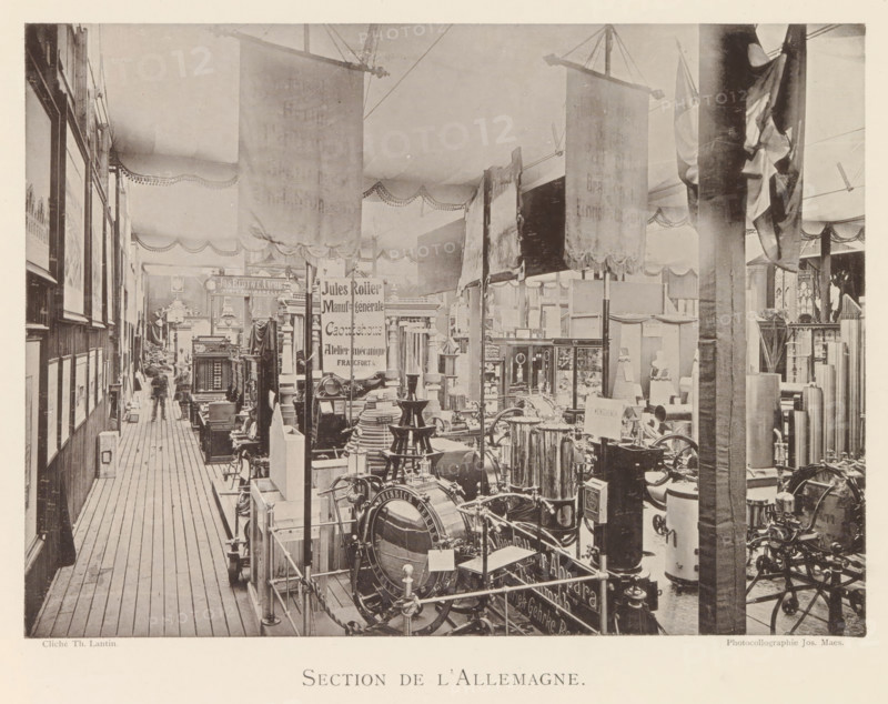 Section de l'Allemagne, Exhibition of German objects during the world exhibition in Antwerp, page, photomechanical print, photographer: Th. Lantin, Antwerp, cliché maker: Joseph Maes, 1894, Th. Lantin, Joseph Maes, photographer, cliché maker, paper, collotype, light printing, height 160 mm x width 220 mm, world fair, world exhibition, exhibition, show, A long indoor exhibition aisle with wooden plank flooring bordered by booths and displays; hanging fabric banners drape from the ceiling and decorative scalloped valances run along the upper walls; rows of framed pictures line the left wall while a variety of machinery, metal equipment, stacked parts, and display cabinets fill the central and right floor space; tall vertical posts and partitions separate sections and a few indistinct human figures are visible near the far end of the aisle; the overall image has a sepia tone and a high vantage capturing depth down the corridor.