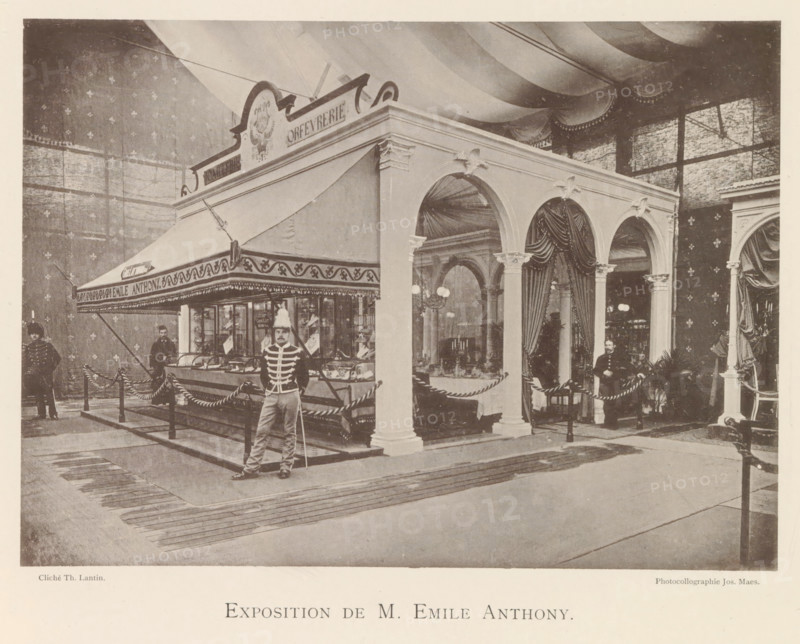 Exposition de M. Emile Anthony, Expositie van de goudsmid Emile Anthony tijdens de wereldtentoonstelling te Antwerpen, page, photomechanical print, photographer: Th. Lantin, Antwerp, clichémaker: Joseph Maes, 1894, paper, collotype, light print, height 160 mm x width 220 mm, jeweler, goldsmith, silversmith, world fair, world exhibition, exhibition, show, A large indoor scene shows a decorative pavilion with arched openings, fluted columns with ornate capitals, and draped curtains inside the arches. A striped canopy extends from one side of the structure above a glass display counter filled with objects, and a rope barrier encircles the area. Several men in uniforms stand near the pavilion, one in front holding a long pole and another seated near the entrance. The background features patterned walls and a high ceiling with sweeping fabric drapery, and there are lamps and furniture visible through the arches. The floor in front of the pavilion has rectangular matting leading toward the s...