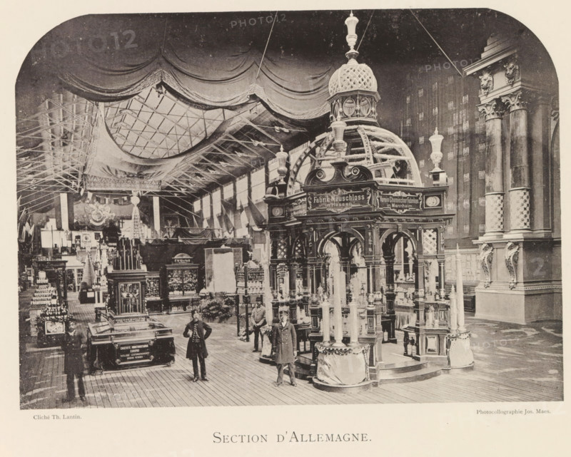 Exhibition of German objects during the World Exhibition in Antwerp, Section d'Allemagne, page, photomechanical print, photographer: Th. Lantin, Antwerp, clichémaker: Joseph Maes, 1894, Th. Lantin, Joseph Maes, photographer, clichémaker, paper, collotype, photoprint, height 158 mm x width 219 mm, world fair, world exhibition, exhibition, show, A large interior exhibition space with a high ceiling draped in fabric and a grid of skylights, ornate display structures with domed and arched tops, tall decorative columns with carved capitals and animal head sculptures, numerous display cases and shelving units filled with objects, freestanding decorative pedestals and urns, several men in period coats standing on a wooden plank floor, hanging flags and banners along the walls, and elaborate decorative moldings and architectural details throughout the hall.