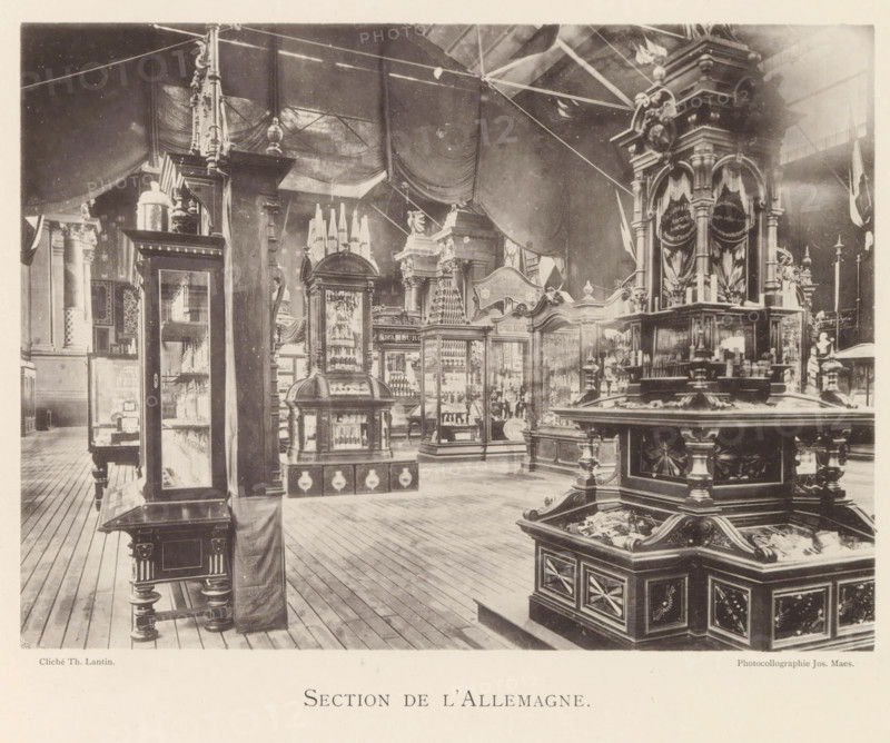 Exhibition of German objects during the world exhibition in Antwerp, Section de l'Allemagne, page, photomechanical print, photographer: Th. Lantin, Antwerp, cliché maker: Joseph Maes, 1894, Th. Lantin, Joseph Maes, photographer, cliché maker, paper, collotype, light printing, height 161 mm x width 218 mm, world fair, world exhibition, exhibition, show, A sepia-toned interior scene showing a large exhibition room with wooden plank flooring and high draped fabric hanging from the ceiling, filled with ornate display cabinets and sculptural wooden fixtures. Prominent carved wooden pedestals and glass-fronted cases stand throughout the space, many with decorative moldings, columns, and finials. A large multi-tiered central display with turned legs and carved panels occupies the foreground, its upper surface holding small objects and decorative elements. In the midground several tall glass vitrines showcase grouped items and have elaborate crowned tops, while a rounded cabinet with a domed t...