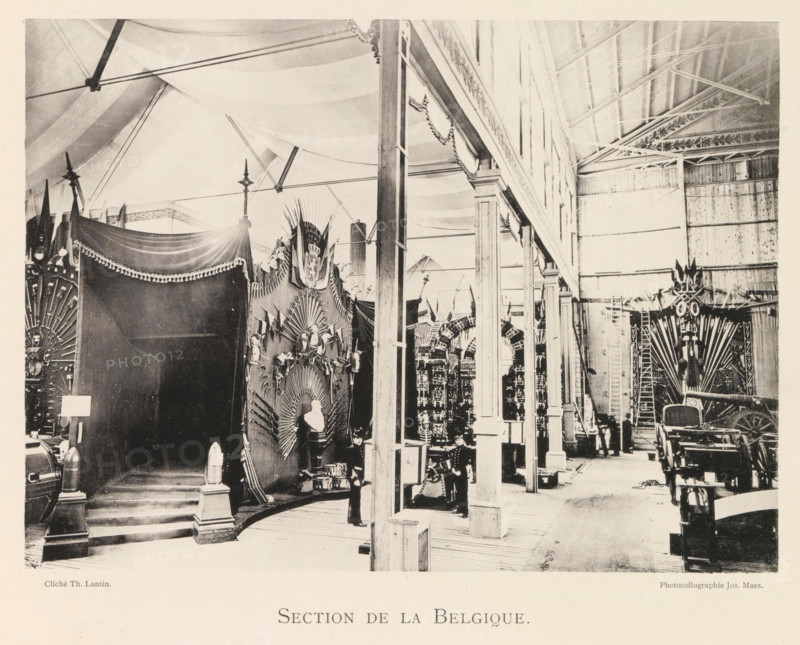 Section de la Belgique, Exhibition of Belgian weapons during the World Exhibition in Antwerp, page, photomechanical print, photographer: Th. Lantin, Antwerp, clichémaker: Joseph Maes, 1894, paper, collotype, photoprint, height 159 mm x width 219 mm, weapons, world fair, world exhibition, exhibition, show, A large indoor space with a high ceiling supported by visible beams and arched fabric drapery stretches across the top, with a row of tall columns and open archways running down the middle right, creating a corridor lined with decorative displays. On the left, a dark draped canopy with fringed trim covers a raised platform with steps leading up to it, flanked by ornamental posts topped with small finials and cylindrical objects on pedestals. Adjacent to the canopy, a tall ornate backdrop features radiating decorative elements, sculpted figures, and a bust on a pedestal, with a uniformed figure standing nearby. Further along the corridor, additional decorative panels and arch-like stru...
