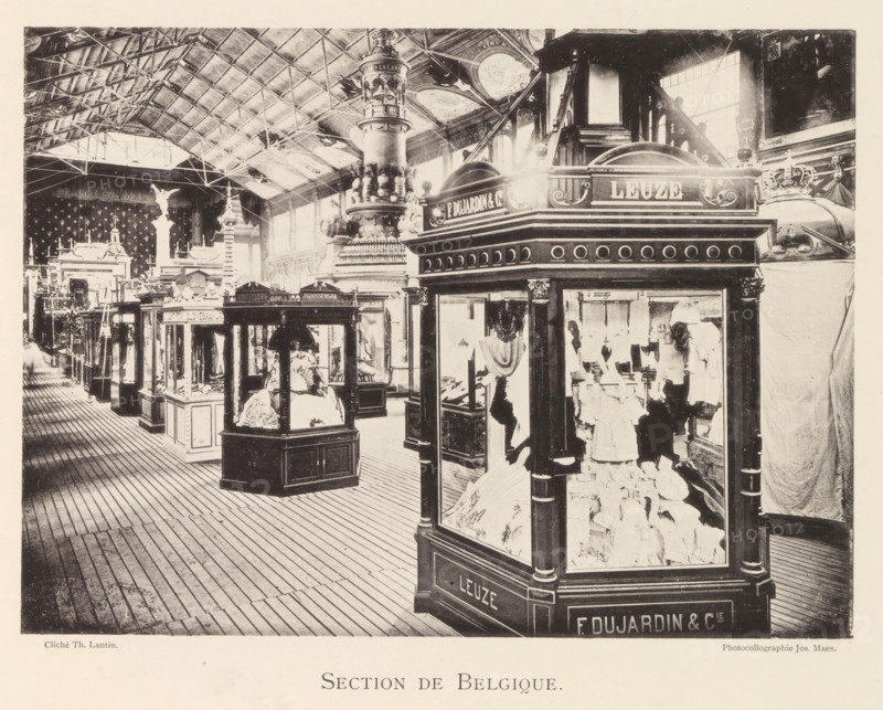 Section de Belgique, Expositie van Belgische beelden tijdens de wereldtentoonstelling te Antwerpen, page, photomechanical print, photographer: Th. Lantin, Antwerp, clichémaker: Joseph Maes, 1894, paper, collotype, light print, height 160 mm x width 220 mm, world fair, world exhibition, exhibition, show, A large indoor exhibition space with a high vaulted ceiling supported by visible metal trusses and skylights, filled with ornate display kiosks and glass cases showcasing garments and decorative objects, a wooden plank floor running parallel to the display rows, an elaborately carved central column with layered decorative elements rising toward the ceiling, arched windows along the far wall letting in light, sculptural figures and ornamental architectural details visible behind and between the kiosks, and various draped fabrics and mannequins positioned within the front glass case.