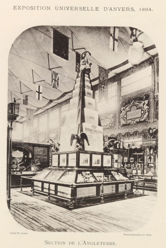 Exhibition of British objects during the World Exhibition in Antwerp, Section de l'Angleterre, page, photomechanical print, photographer: Th. Lantin, Antwerp, clichémaker: Joseph Maes, 1894, Th. Lantin, Joseph Maes, photographer, clichémaker, paper, collotype, photoprint, height 220 mm x width 160 mm, world fair, world exhibition, exhibition, show, A large indoor exhibition hall with high windows and decorative wall panels, featuring a central tall pyramid-like display topped by a seated female figure and flanked by sculpted eagle figures; glass-topped wooden display cases surround the base of the central structure, containing objects and framed panels on their sides; flags and bunting hang from the ceiling across the space; ornate signage and decorative motifs adorn the walls behind the displays; wooden floorboards run across the foreground.