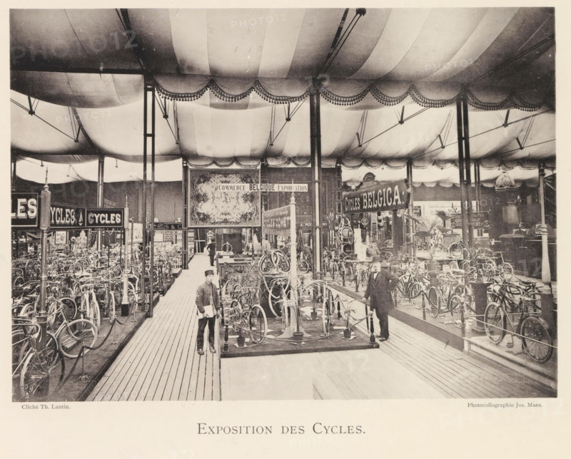 Exposition des Cycles, Expositie van fietsen tijdens de wereldtentoonstelling te Antwerpen, page, photomechanical print, photographer: Th. Lantin, Antwerp, clichémaker: Joseph Maes, 1894, paper, collotype, light print, height 157 mm x width 220 mm, bicycle, world fair, world exhibition, exhibition, show, A large indoor exhibition space filled with rows of bicycles displayed on stands beneath a fabric ceiling with scalloped valances and decorative fringe, supported by tall slender columns and overhead rigging; wooden plank walkways run between the displays and a few men in period clothing stand among the bicycles, one near the foreground holding papers and another leaning on a rail, while ornate signs and framed panels hang above the exhibits and potted plants are placed near some of the displays.