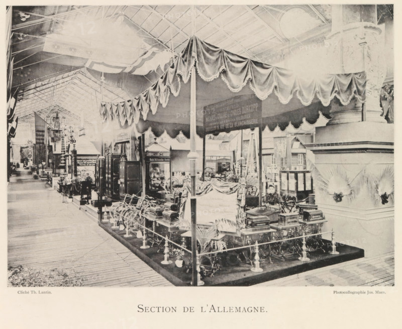 Exhibition of German objects during the world exhibition in Antwerp, Section de l'Allemagne, page, photomechanical print, photographer: Th. Lantin, Antwerp, cliché maker: Joseph Maes, 1894, Th. Lantin, Joseph Maes, photographer, cliché maker, paper, collotype, photoprint, height 158 mm x width 220 mm, world fair, world exhibition, exhibition, show, A large indoor exhibition space with a high vaulted roof and visible metal trusses, rows of display booths and ornate decorative columns, a central raised platform area enclosed by rope and stanchions holding various ornate machinery and wooden cabinets, a fabric canopy with scalloped and fringed drapery suspended over the central area supported by slender poles, decorative plants placed around the displays, a person seated near the front of the exhibit partially visible behind equipment, additional booths and objects extending into the distance along a wooden plank floor, and decorative architectural elements including carved column bases a...