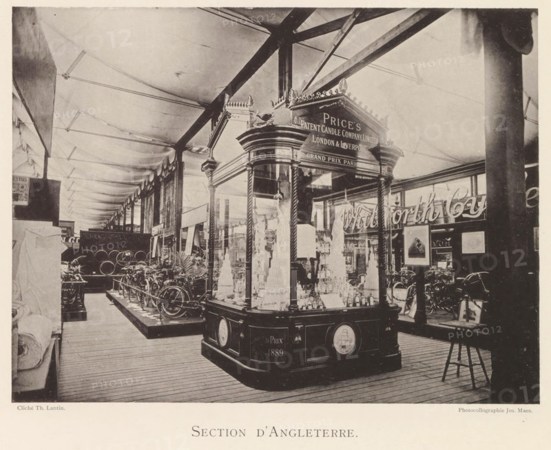 Section d'Angleterre, Exhibition of British objects during the World Exhibition in Antwerp, page, photomechanical print, photographer: Th. Lantin, Antwerp, clichémaker: Joseph Maes, 1894, Th. Lantin, Joseph Maes, photographer, clichémaker, paper, collotype, photoprint, height 160 mm x width 214 mm, world fair, world exhibition, exhibition, show, A large indoor exhibition space with a high fabric ceiling supported by visible beams and columns, showing a decorative wooden display pavilion with glass panels and ornate trim in the foreground filled with tiered objects and bottles, flanked by rope-twisted columns and a carved cornice topped by ornamental finials. Around the pavilion are rows of bicycles and metal wheels arranged on raised platforms, various framed pictures and signs mounted on walls and stands, potted plants near the floor, wooden plank flooring underfoot, and a tall dark column partially obscuring the right side of the scene. The image has a sepia tone and shows detailed s...