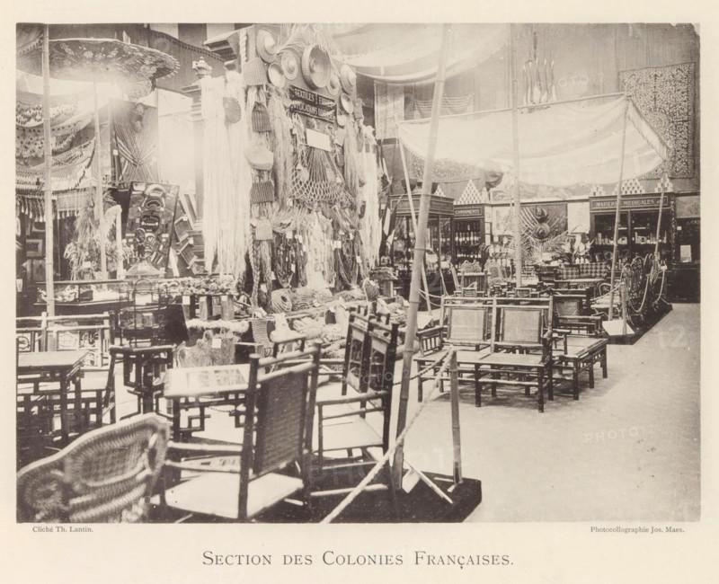 Section des Colonies Françaises, Exhibition of the French colonies during the World Exhibition in Antwerp, page, photomechanical print, photographer: Th. Lantin, Antwerp, cliché maker: Joseph Maes, 1894, paper, collotype, photoprint, height 160 mm x width 219 mm, colonial system, world fair, world exhibition, exhibition, show, A large indoor scene showing many wooden chairs arranged in rows and stacks across the foreground and midground, woven seats and backs visible on several pieces, with a variety of baskets, ropes, and hanging textile fringes displayed on a central vertical structure. Canopy fabrics and draped cloth hang from poles overhead, and decorative plates and woven mats are mounted on the display. Shelving units and display cabinets filled with bottles and containers sit toward the rear, flanked by patterned wall hangings and carved panels. Several poles and ropes run through the space, and small tables, stools, and ornamental objects are scattered among the furniture and d...
