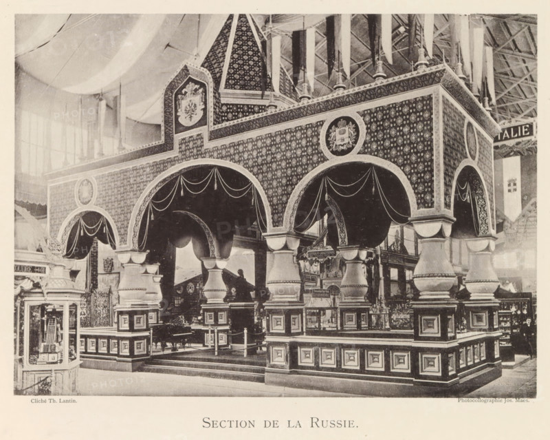 Section de la Russie, Exhibition of Russian objects during the World Exhibition in Antwerp, page, photomechanical print, photographer: Th. Lantin, Antwerp, clichémaker: Joseph Maes, 1894, paper, collotype, photoprint, height 160 mm x width 219 mm, world fair, world exhibition, exhibition, show, A large ornate pavilion with patterned walls and decorative arches stands inside a spacious hall; the structure features rounded archways draped with fabric swags, heavy column-like supports with layered bases and capitals, and panels with repeating motifs covering the upper surfaces. Above the arches a peaked roof section with patterned surfaces and circular medallions bearing emblem-like designs rises toward the ceiling, where hanging fabric banners descend from an exposed truss roof. In front of and beneath the arches are display areas and small stands with varied objects, and a framed glass cabinet sits at one side showing items inside. The scene is monochrome and shows varied textures of ca...