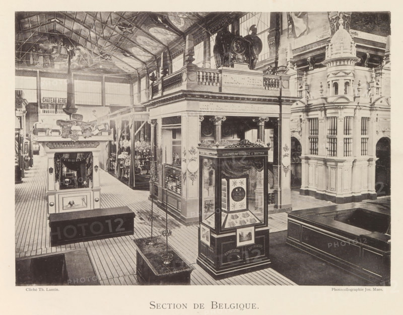 Section de Belgique, Expositie van Belgische beelden tijdens de wereldtentoonstelling te Antwerpen, page, photomechanical print, photographer: Th. Lantin, Antwerp, clichémaker: Joseph Maes, 1894, paper, collotype, light print, height 160 mm x width 220 mm, world fair, world exhibition, exhibition, show, A sepia-toned photograph showing an indoor exhibition hall with high vaulted ceiling and visible rafters, a wide wooden plank floor, and tall windows along the back wall; ornate multi-level architectural displays and pavilions fill the space, featuring classical columns, balustrades, carved decorative elements, and a domed turret-like structure on the right; a freestanding glass display case with decorative panels and framed images stands near the foreground, and a smaller kiosk with sculpted ornamentation and draped fabric is visible toward the left; hanging chandeliers and tall lampstands are arranged along the center aisle, and carved statues and reliefs decorate the upper terraces a...