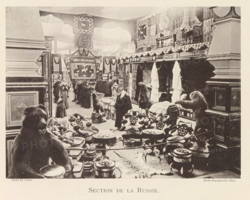 Section de la Russie, Exhibition of Russian fur products during the world exhibition in Antwerp, page, photomechanical print, photographer: Th. Lantin, Antwerp, cliché maker: Joseph Maes, 1894, paper, collotype, photoprint, height 160 mm x width 220 mm, spoils of the hunt, game, venison, world fair, world exhibition, exhibition, show, A large indoor scene shows an exhibition-like space filled with animal hides, fur garments, and stuffed animal figures arranged on platforms and tables. In the foreground a seated bear figure faces slightly toward the viewer with a bowl in front of it, surrounded by low round stools topped with patterned cushions and small carved objects. Further back there are more furry seated figures and standing animal forms placed on draped surfaces and rugs, with rows of hanging coats and long fur garments visible along the midground. The walls and partitions are decorated with large patterned textiles and banners, and sculpted animal heads and pelts are displayed o...