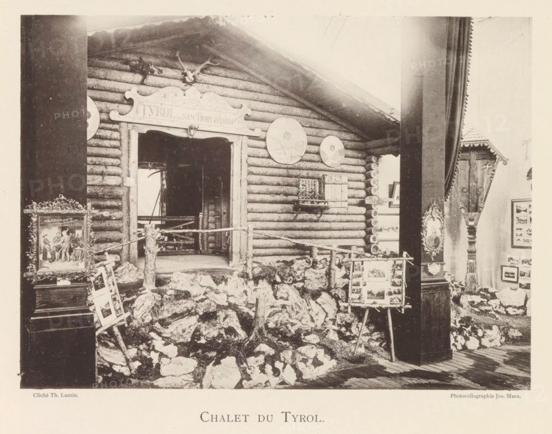 Chalet du Tyrol, Tyrolean chalet in the world exhibition in Antwerp, page, photomechanical print, photographer: Th. Lantin, Antwerp, cliché maker: Joseph Maes, 1894, paper, collotype, light printing, height 161 mm x width 219 mm, world fair, world exhibition, exhibition, show, log-cabin, A photograph of a rustic log facade with a peaked roof and a doorway framed by carved wood, mounted antlers above the entrance, circular plaques on the exterior wall, a small shuttered window with a decorative grille, and a rough rocky foreground with a simple wooden fence; an easel displaying mounted images stands among the rocks, an ornate framed painting rests nearby, and a tall carved column with decorative panels and a small roofed shrine is visible to the right, all within an interior space with wooden floorboards and vertical supporting beams.